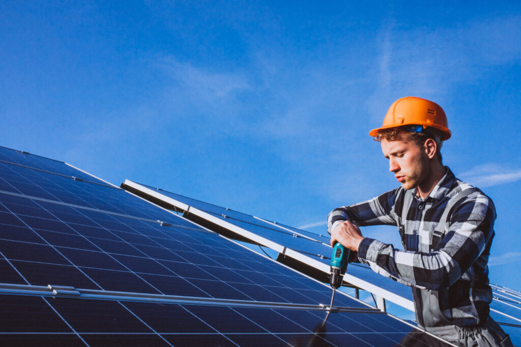 Man in orange hard hat installing solar panels with a power drill against a clear blue sky, illustrating electrical safety and renewable energy installation.